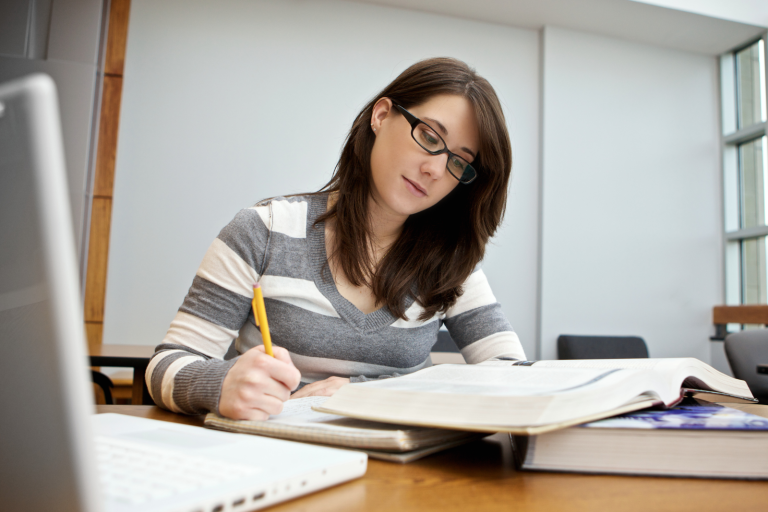 Student writing notes at a desk with a laptop and textbooks in a study space