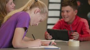 Still from accessible texts video showing one female student writing on paper and a male student in the background holding an ipad