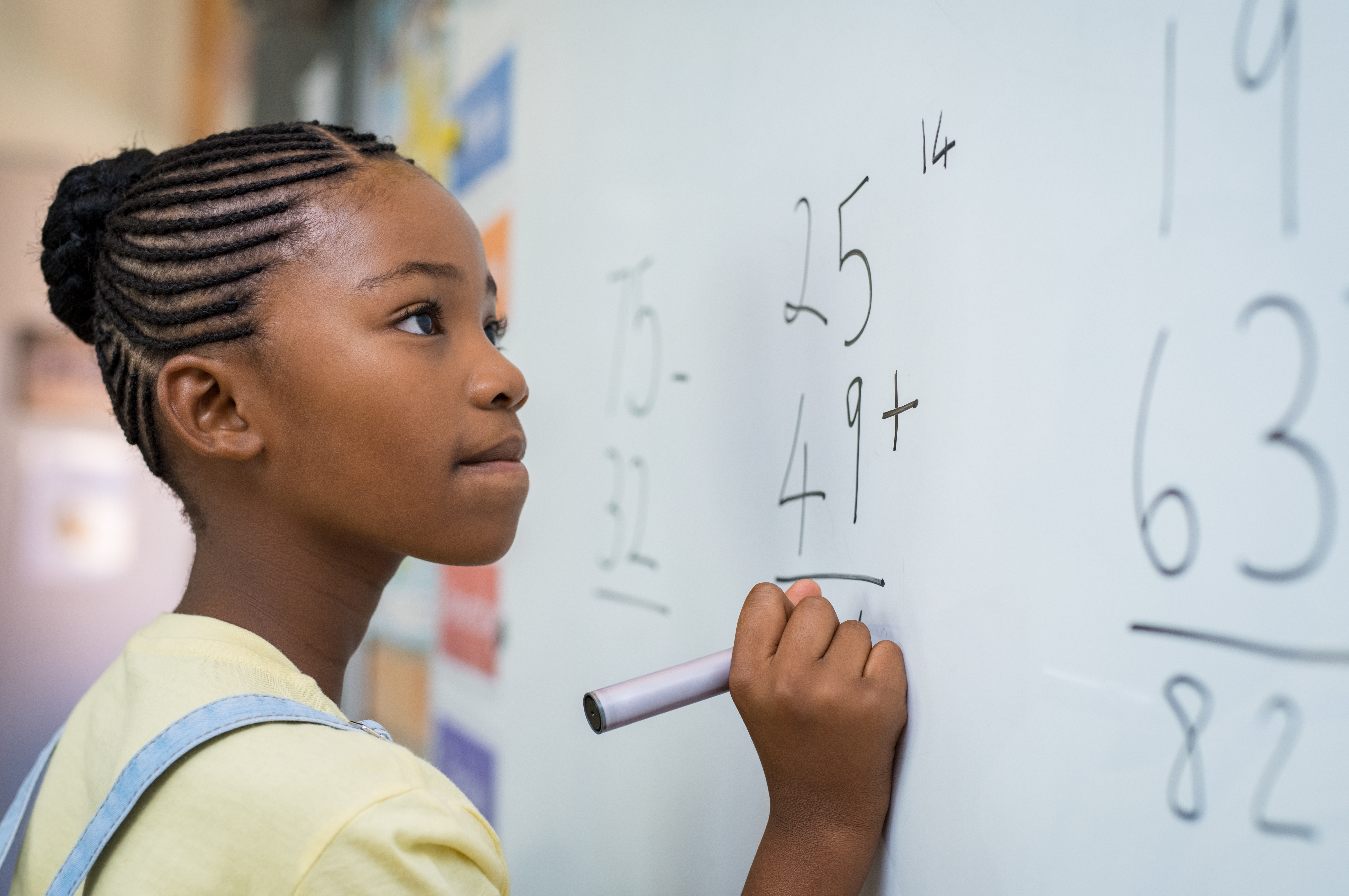 Young girl stands at white board with marker in hand, pondering a math equation.