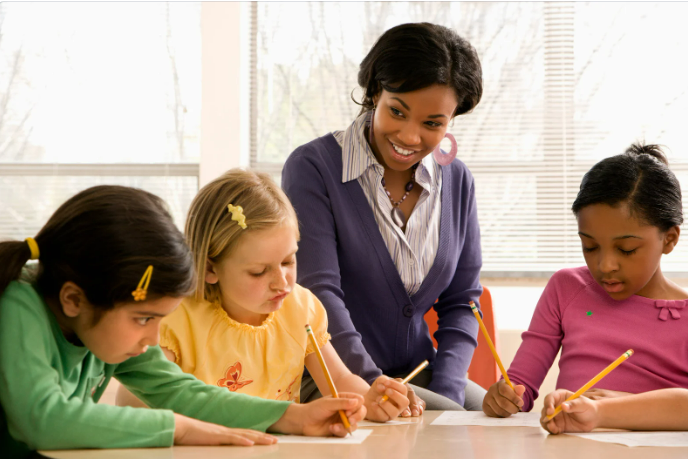 Elementary teacher guiding a small group of students as they complete a structured writing activity at a classroom table.